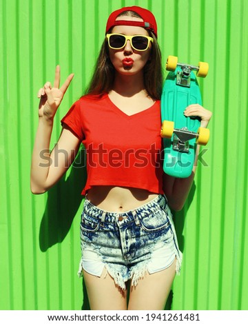 Summer portrait of young woman with green skateboard wearing a shorts and red baseball cap in a city