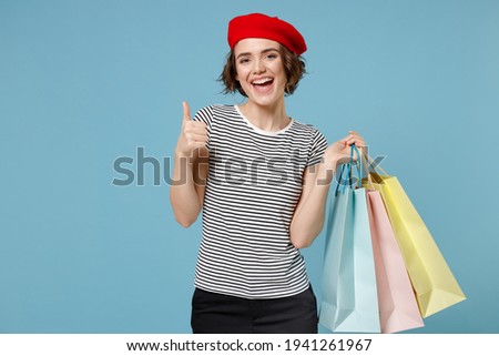 Young happy woman 20s with short hairdo in french beret red hat striped t-shirt hold package bags with purchases enjoy shopping show thumb up gesture isolated on pastel blue background studio portrait