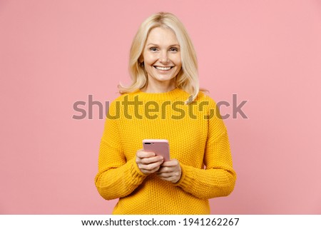 Smiling elderly gray-haired blonde woman lady 40s 50s years old in yellow casual sweater standing using mobile cell phone typing sms message isolated on pastel pink color background studio portrait Royalty-Free Stock Photo #1941262267