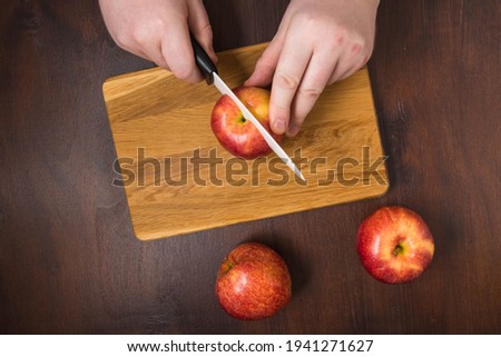 The dark food photography of chopping board and red apples on kitchen tabletop. The flat lay view of cook hands with white knife and red apples on the brown wooden table. The cook is cutting fruits.