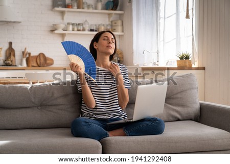 Tired millennial woman suffers from stuffiness and an inoperative air conditioner, waving blue fan sitting on couch at home working on laptop computer. Overheating high temperature, hot summer weather Royalty-Free Stock Photo #1941292408
