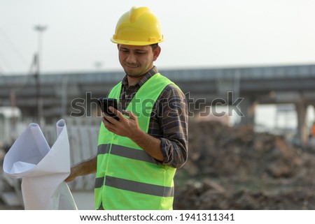 Asian construction workers use smartphone and hold a plan blueprint at the construction site.