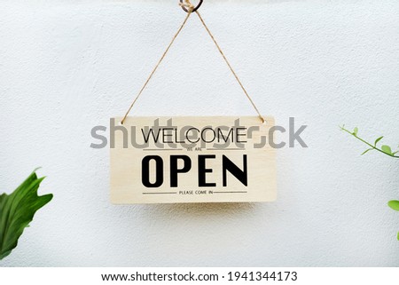 welcome we're open. black text on wood sign hanging on white wall  hanve plant in forground