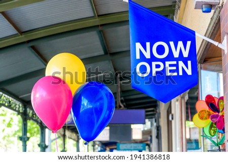 A blue sign with the words NOW OPEN is hanging next to three balloons to advertise an open business