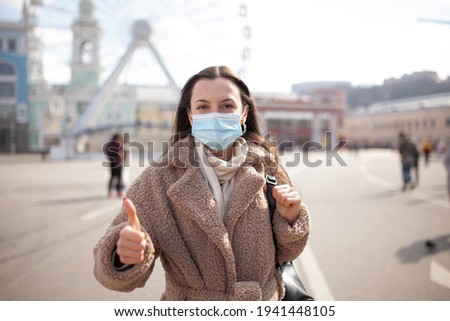Woman doing thumbs up gesture wearing a protective mask. Young woman in medical sterile protective mask on her face, walking in a European city, showing thumbs up
