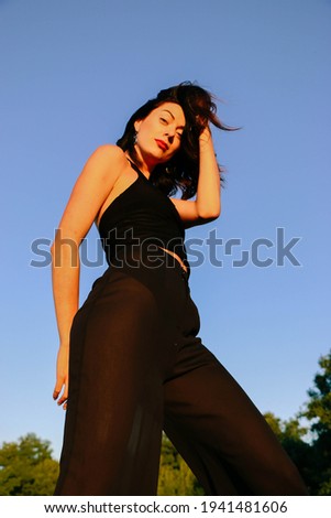 Young beautiful brunette in fashionable black clothes posing on a sunny summer evening, sunset light. Attractive woman happily dancing, posing against blue sky background