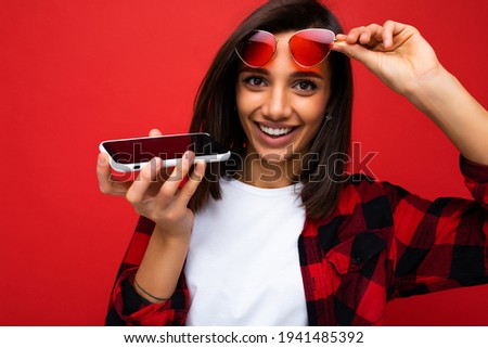 photo of beautiful smiling happy young brunet woman wearing stylish red shirt white t-shirt and red sunglasses isolated over red background using mobile phone recording voice message looking at camera