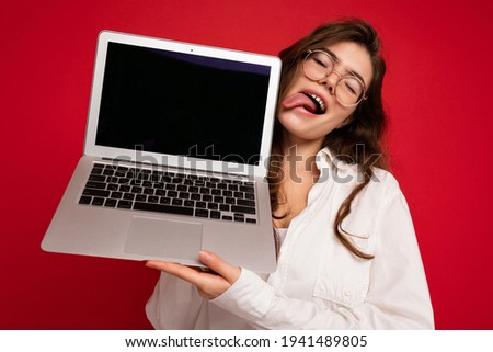 Crazy mad beautiful brunette curly young woman holding computer laptop wearing glasses white shirt showing tongue looking to the side isolated over red wall background
