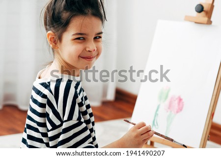 Candid portrait of a little girl kid smiling, looking at the camera, and painting on the paper on an easel at home. A pretty kid sitting on the carpet and drawing in her room.