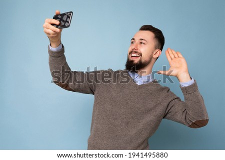 Handsome young man wearing casual stylish clothes standing isolated over background wall holding smartphone taking selfie photo looking at mobile phone screen display showing hand palm saying hello