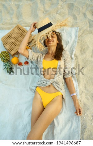 joyful happy woman in yellow bikini lying on blanket at beach next to beach accessories and fruit