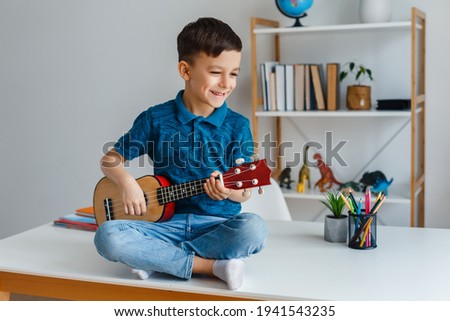 Talented kid playing soprano ukulele sitting on desk. Preschool boy learning guitar at leisure. Concept of early childhood education and music hobby Royalty-Free Stock Photo #1941543235