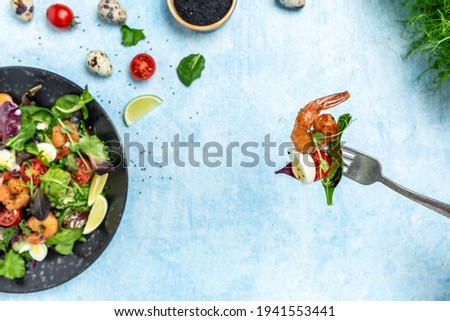 Fork with salad from vegetables cherry tomatoes, cucumber, avocado, eggs and smoked shrimps. Delicious breakfast or snack on a blue background, top view.