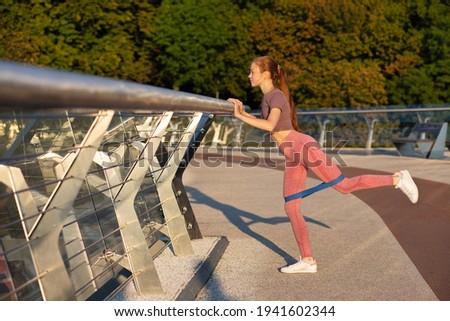 Athletic red haired woman wears sport t shirt and leggins exercising with a rubber band at the bridge. Space for text

