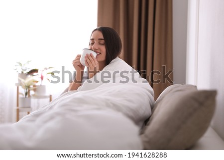 Woman covered in blanket drinking cup of hot beverage on sofa