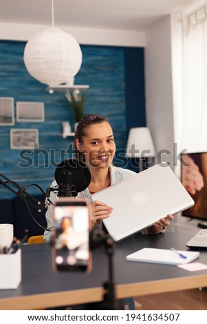 Influencer presenting new laptop for giveaway during podcast. Creative content creator making video blog concept speaking and looking at smartphone on tripod at podcast home studio broadcast