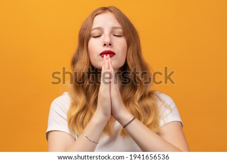 Pretty girl with wavy redhead, wearing white t-shirt holding palms together. She meditating, praying for peace and love, having calm and peaceful facial expression. Isolated on yellow background