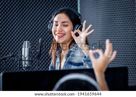 female studio technician of the computer monitor of Software for recording in foreground of young Asian woman vocalist Wearing Headphones recording a song front of microphone in a professional studio