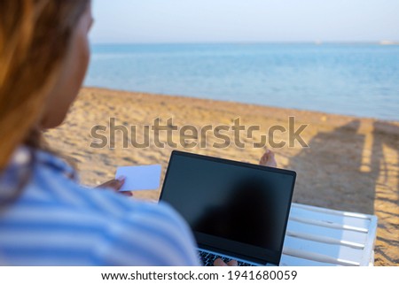 Mockup image of a woman using laptop with blank desktop screen and holds credit card while sitting by the sea with blue sky background. Freelance work concept. Technology and travel.
