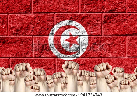 Raised fists against the background of the depicted flag of Tunisia on a brick wall, a concept of strength and unity.
