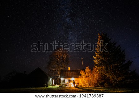 Milky way with stars in clear night. Old country house with burning fire outdoors.