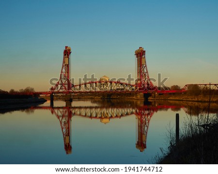 Image shows the Newport Bridge across the centre mid-ground of the image against a gradated blue and orange sky. The foreground shows a the River Tees reflecting the scene and sky. Royalty-Free Stock Photo #1941744712