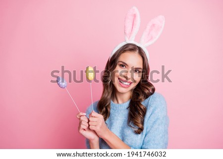 Photo of lady hold two painted eggs on sticks beaming smile wear rabbit ears headband blue sweater isolated pink background