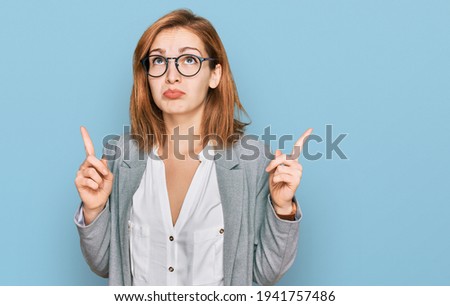 Young caucasian woman wearing business style and glasses pointing up looking sad and upset, indicating direction with fingers, unhappy and depressed. 