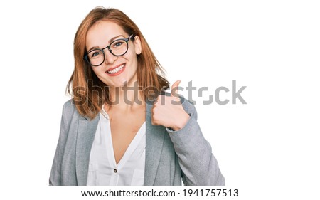 Young caucasian woman wearing business style and glasses doing happy thumbs up gesture with hand. approving expression looking at the camera showing success. 