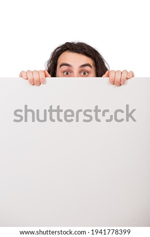 Fearful man lurking behind a blank whiteboard. Empty sheet with copy space for advertising and messages. Timid introvert guy hiding identity behind a white paper banner. Young male looking surprised