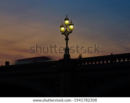 Low-key, soft image of a gothic-style streetlight on Westminster Bridge, flanked by a silhouetted and motion-blurred London Bus and person ahead of the dying embers of a summer sunset. 
