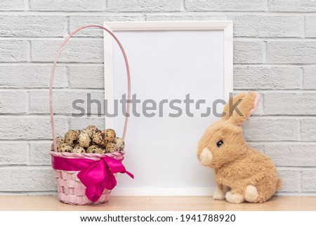 Close-up of basket full of easter eggs, fluffy bunny toy, empty white photo frame. Cute home interior decoration. Happy easter holiday, spring season concept, copy space