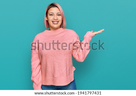 Hispanic woman with pink hair wearing casual winter sweater smiling cheerful presenting and pointing with palm of hand looking at the camera. 
