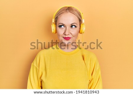 Young blonde woman listening to music using headphones smiling looking to the side and staring away thinking. 