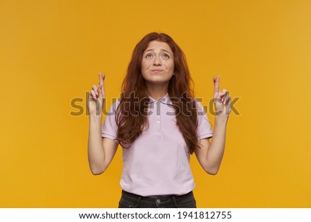 Cute, positive woman with long ginger hair. Wearing pink t-shirt. People and emotion concept. Keeps fingers crossed, making a wish. Watching up at copy space, isolated over orange background