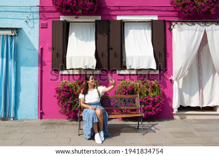 A young tourist woman with a backpack looks at her mobile in the colorful streets of Burano in Venice while smiling and visiting the city, Italy