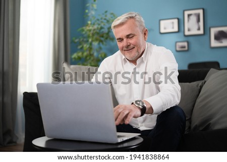 Smiling happy middle aged 50s old senior man using laptop computer technology working online, surfing internet, typing email, shopping in ecommerce store relaxing with device sitting on couch at home.