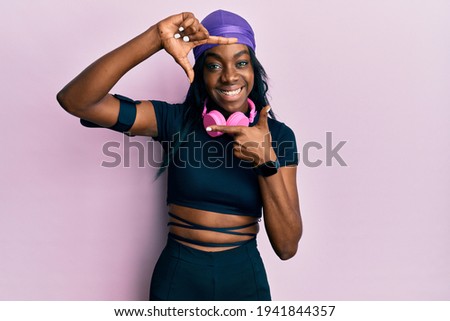 Young african american woman wearing gym clothes and using headphones smiling making frame with hands and fingers with happy face. creativity and photography concept. 