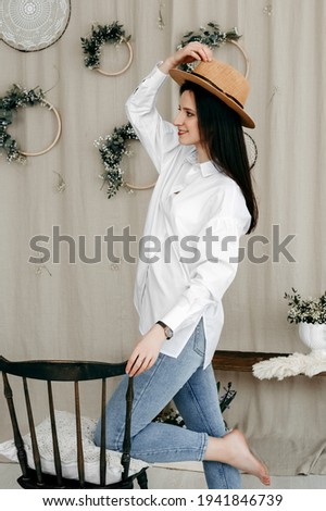 Photo of a beautiful young brunette in a straw hat on her head. She puts her knee on the seat of the wooden chair. Girl in a white shirt and blue jeans.