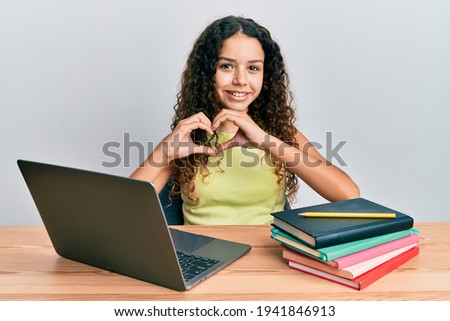 Teenager hispanic girl sitting on the table studying for school smiling in love doing heart symbol shape with hands. romantic concept. 