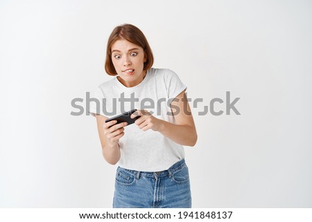Portrait of girl focused on video game, playing on smartphone and biting lip, tilting body with cell phone, white background