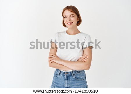 Portrait of beautiful natural female model with short hair, wearing t-shirt with jeans, cross arms on chest and smile at camera with happy emotion, white background