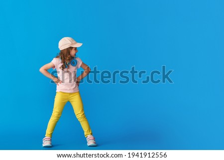 little cute girl in fashionable summer clothes and headphones poses on a blue background in full growth and looks away. The concept of summer and spring casual children's clothing. Copy space