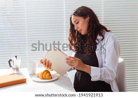 Side view of happy young beautiful woman reading book while having morning breakfast with coffee and croissants on background of blinds. Good morning concept and pleasant lunch break