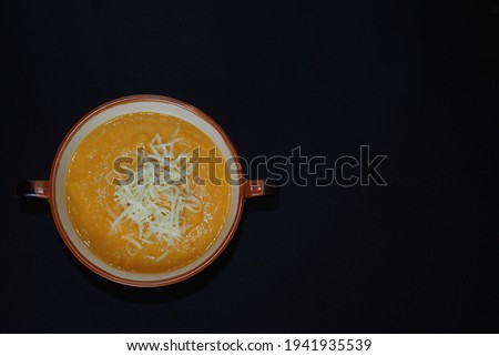 pumpkin and carrot cream soup with grated cheese stands in a round brown plate with handles on a black background top view . dietary home food