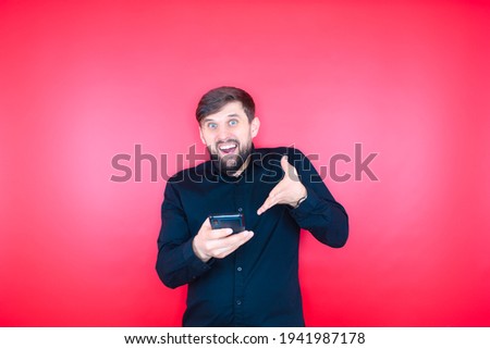 A bearded man in a black shirt holds a phone in his hand and gestures emotionally. Bearded man on a red background