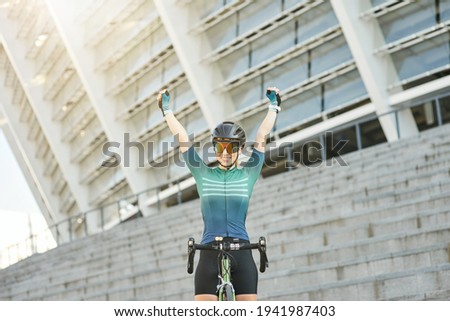 Excited professional female cyclist in cycling garment raised her arms, smiling at camera while standing with her bike outdoors on a daytime