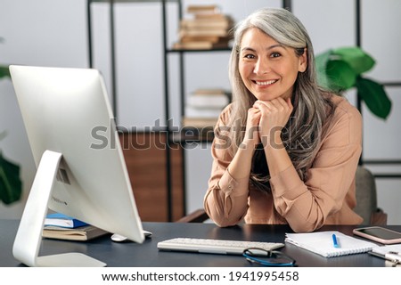 Successful influential attractive middle aged Asian female, business woman, manager, freelancer, in stylish clothes, works at computer, rests her head with her hands, looks at camera, smiles friendly Royalty-Free Stock Photo #1941995458