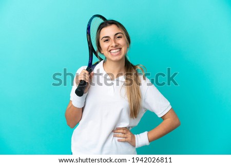 Young woman tennis player isolated on blue background posing with arms at hip and smiling