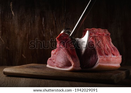 piece of raw pork ribbed loin chopped with an kitchen ax lies on cutting board with brown old wooden backdrop in shadow. side view. artistic moody photo in simple rustic style with copy space